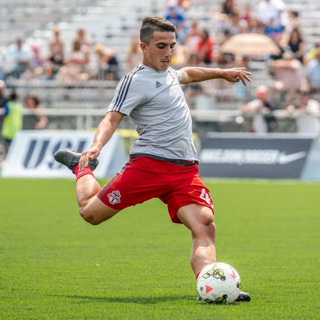 Luca in a grey warm-up top and red shorts striking the ball, a USL banner behind.